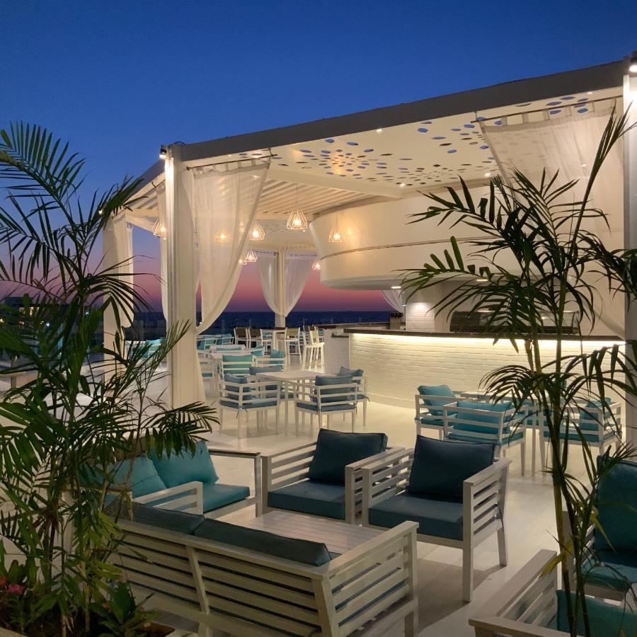 โรงแรมซันไรส์ อเล็กซ์ อเวนิว อเล็กซานเดรีย ภายนอก รูปภาพ The photo shows a stylish outdoor terrace or rooftop lounge area. It features white furniture, including comfortable seating with teal cushions. The space is adorned with draped sheer curtains, providing a relaxed and elegant atmosphere. Lush green p