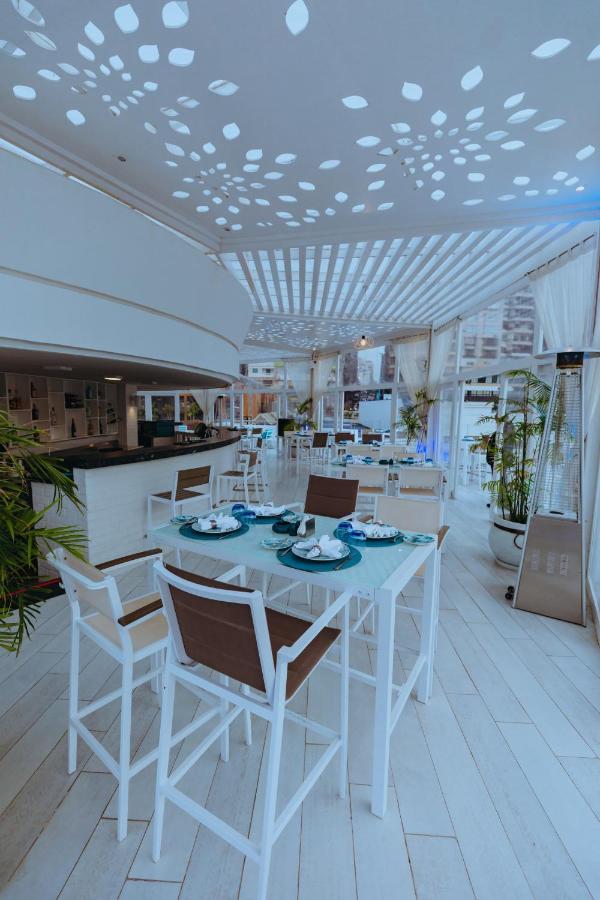 โรงแรมซันไรส์ อเล็กซ์ อเวนิว อเล็กซานเดรีย ภายนอก รูปภาพ The photo shows a bright and modern restaurant interior. The space features white and light-colored furniture, including tables with a combination of white and brown chairs. Each table is elegantly set with plates, utensils, and decorative elements. 