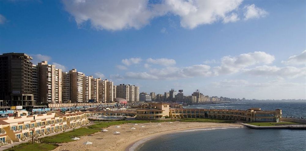โรงแรมซันไรส์ อเล็กซ์ อเวนิว อเล็กซานเดรีย ภายนอก รูปภาพ The photo shows a scenic coastal view featuring a sandy beach and a curved shoreline. In the foreground, there are several sun loungers and a landscaped area. Beyond the beach, there are multiple high-rise buildings lining the waterfront, indicating 