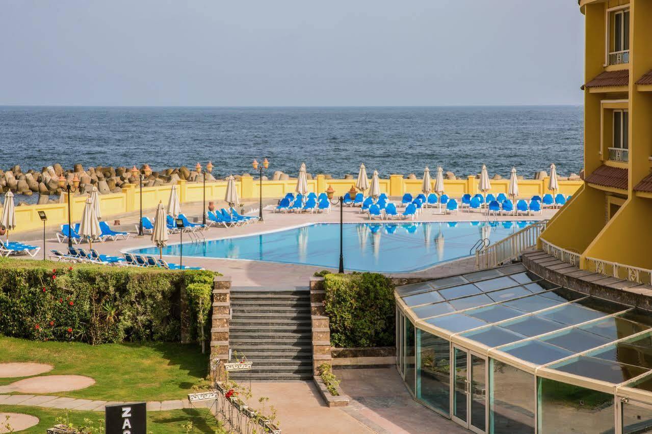 โรงแรมซันไรส์ อเล็กซ์ อเวนิว อเล็กซานเดรีย ภายนอก รูปภาพ The photo depicts a scenic view of a pool area adjacent to the ocean. In the foreground, there is a large swimming pool surrounded by numerous blue lounge chairs and umbrellas, indicating a relaxing setting. There are steps leading down from a grassy